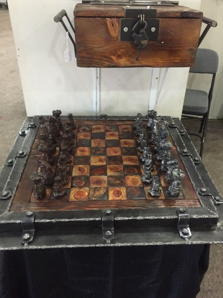 Looking down on a chess board of dark and light squares in a steel frame with strap brackets, on a black-draped table, with a wooden box with a steel latch above it.