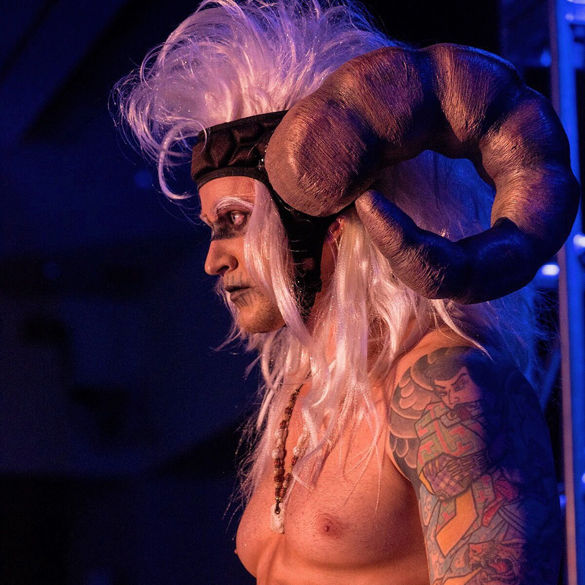 In blue light, a man in profile wears large curling horns on a headband with long white hair.