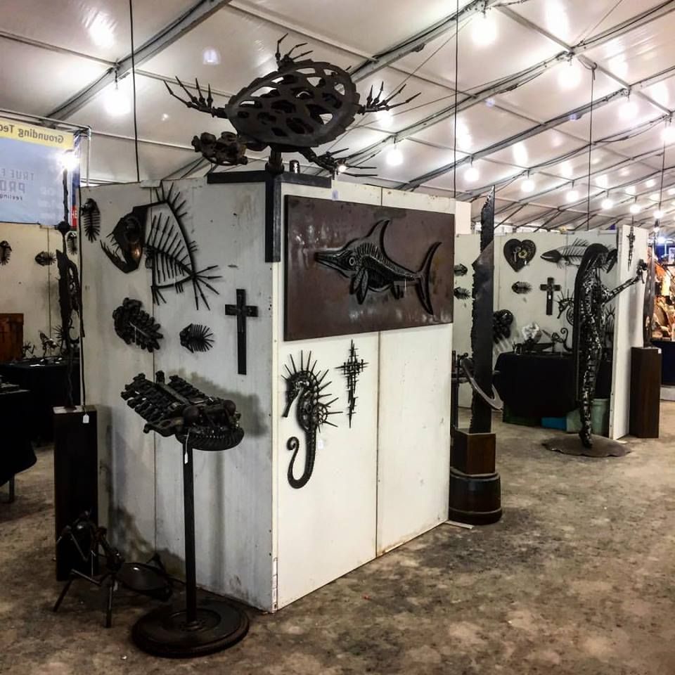 Inside a white show tent, a large steel trilobite sits on top of a white booth wall hung with a fish, crosses and a seahorse.