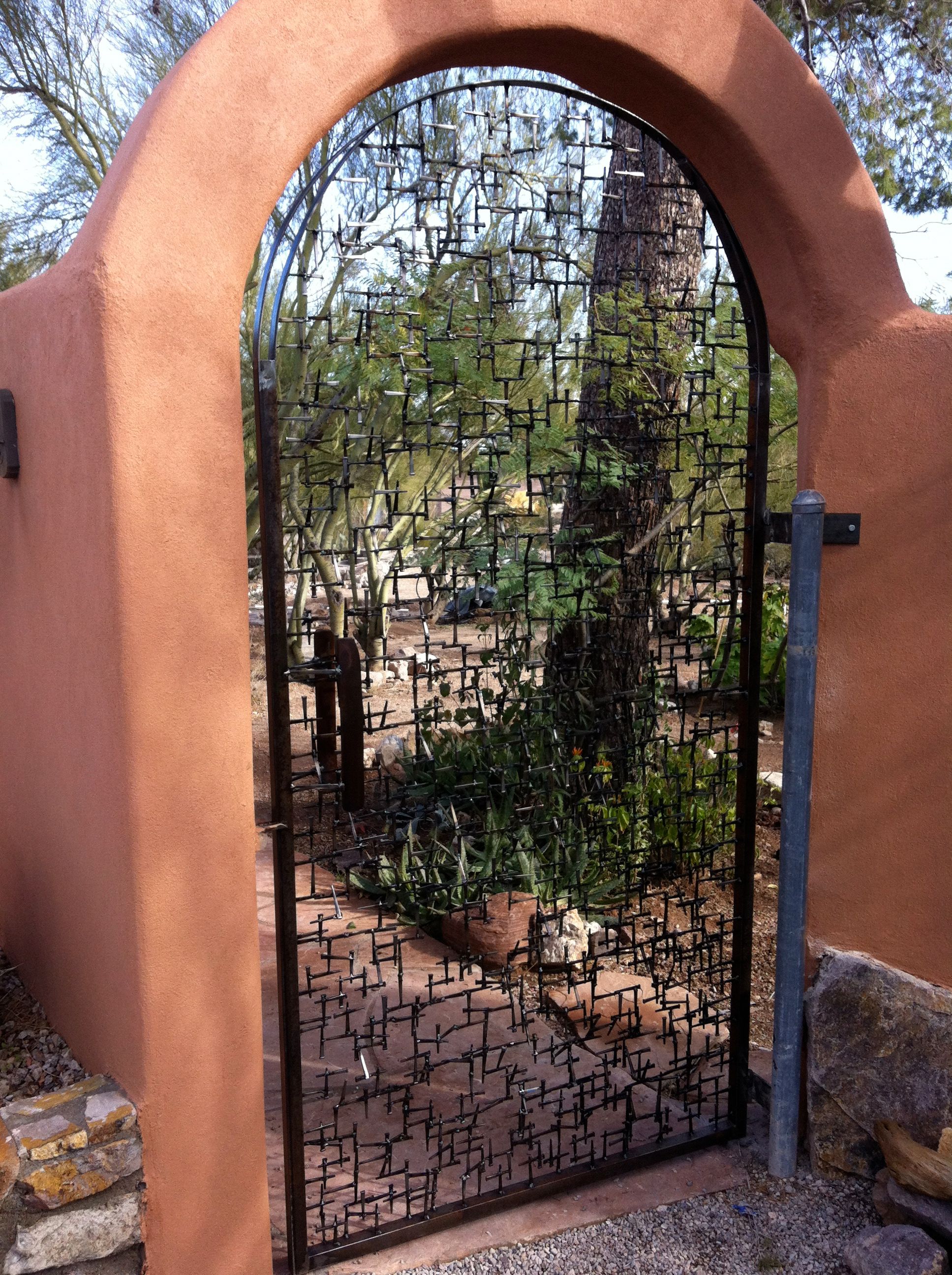 An arched steel gate filled with a lattice of welded nails, set in a rose-colored adobe wall, with a garden path and plants visible through it.