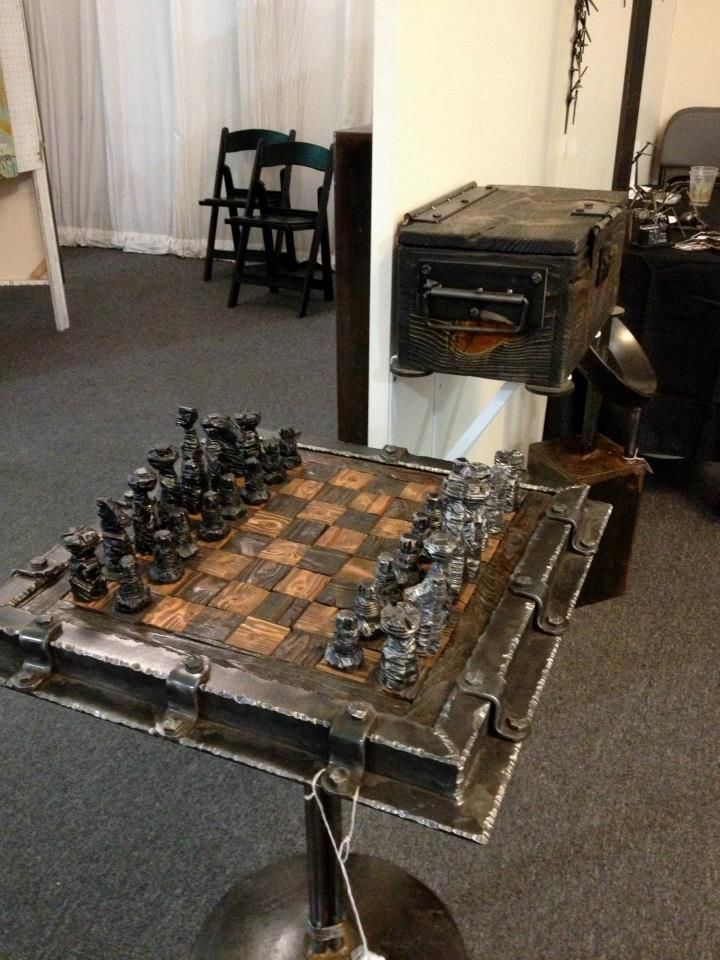 A chess set in a riveted steel frame on a small pedestal stand, seen from one corner, with a wooden box on a shelf behind it.